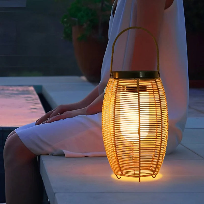 A warm-toned, cylindrical outdoor lantern made of woven material emits a soft glow as it sits on a light-colored ledge next to a person wearing a white dress at dusk or night. The lantern has a dark top, possibly a solar panel, and a handle for carrying or hanging. A dark pool and a potted plant are visible in the blurred background.