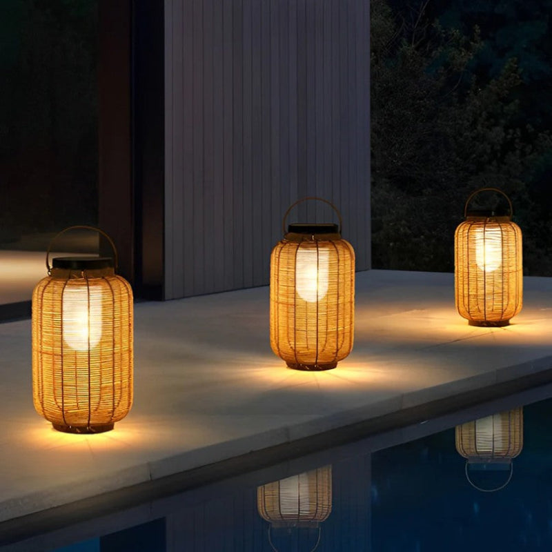 Three warm-toned, cylindrical outdoor lanterns made of woven material emit a soft glow as they sit on a stone surface beside a dark pool at dusk or night. The lanterns have dark tops, possibly solar panels, and metal handles. Their reflections are visible in the water.