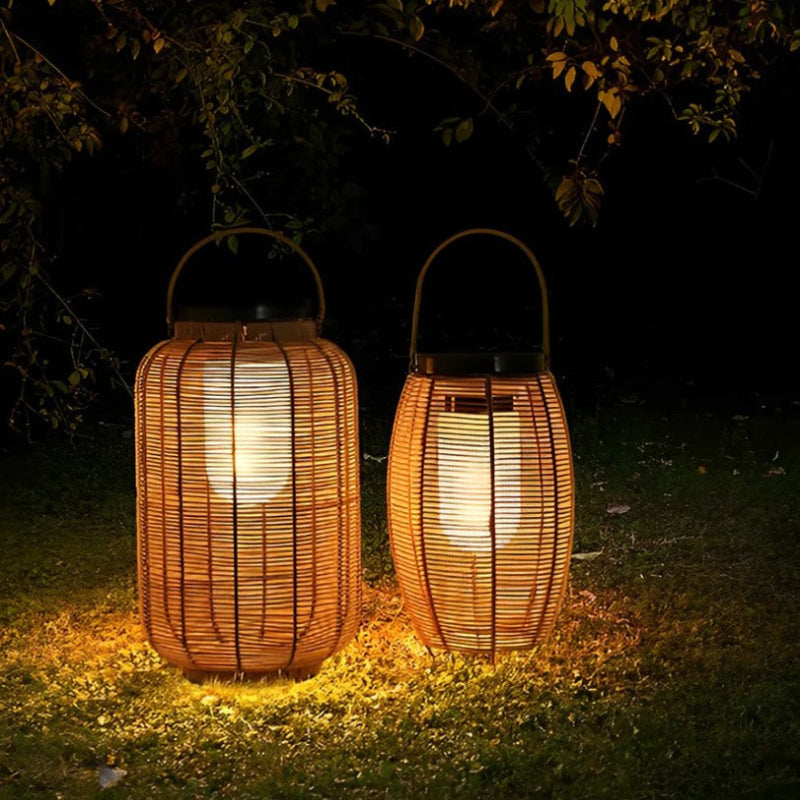 Two warm-toned, cylindrical outdoor lanterns made of woven material emit a soft white glow as they sit on grass at night. The lanterns have dark tops, likely solar panels, and metal handles for hanging. Dark foliage is visible in the background.