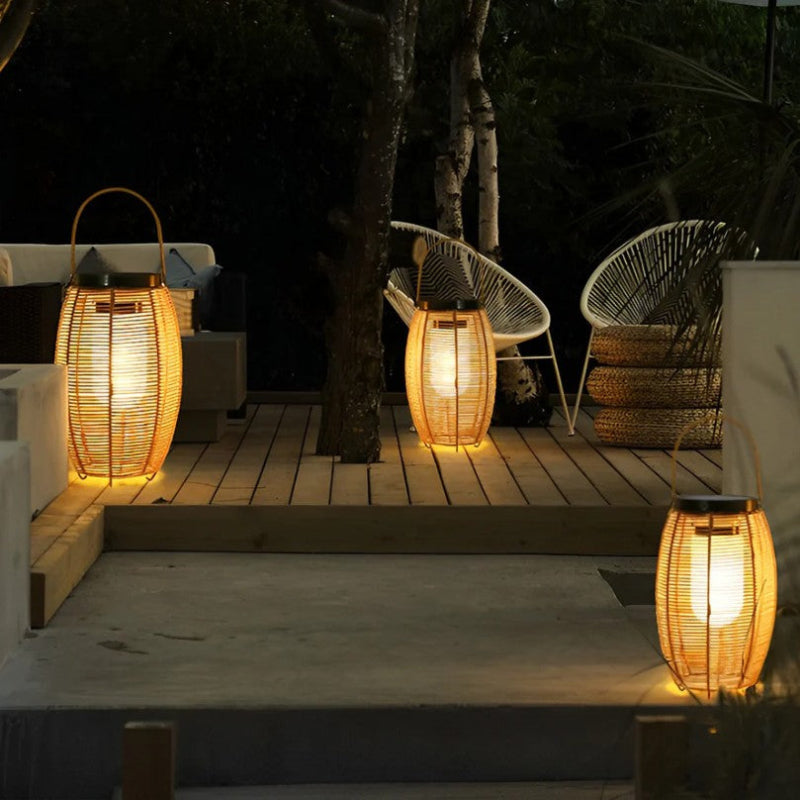 Three warm-toned, cylindrical outdoor lanterns made of woven material emit a soft white glow on a patio at night. The lanterns have dark tops, likely solar panels, and metal handles. Outdoor furniture and trees are visible in the background.