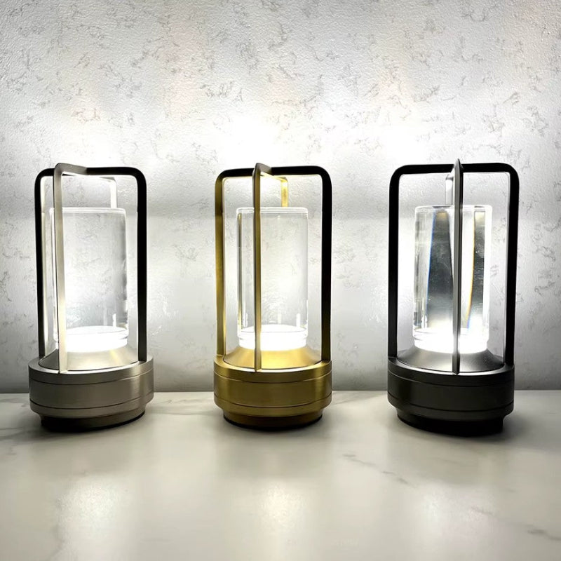 Three small, portable LED table lamps are lined up on a light-colored surface against a textured off-white wall. Each lamp has a cylindrical clear glass diffuser and a metal frame and base in different finishes: gray, gold, and black. The lamps emit a bright, upward glow, illuminating the glass diffusers.