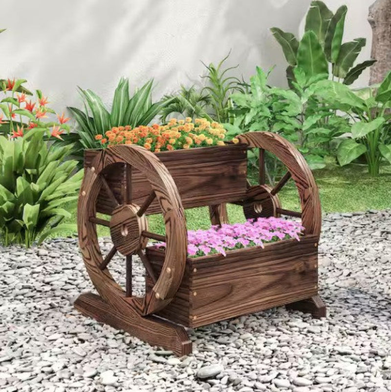 Wooden planter with decorative wheel design on a pebble surface with plants in the background