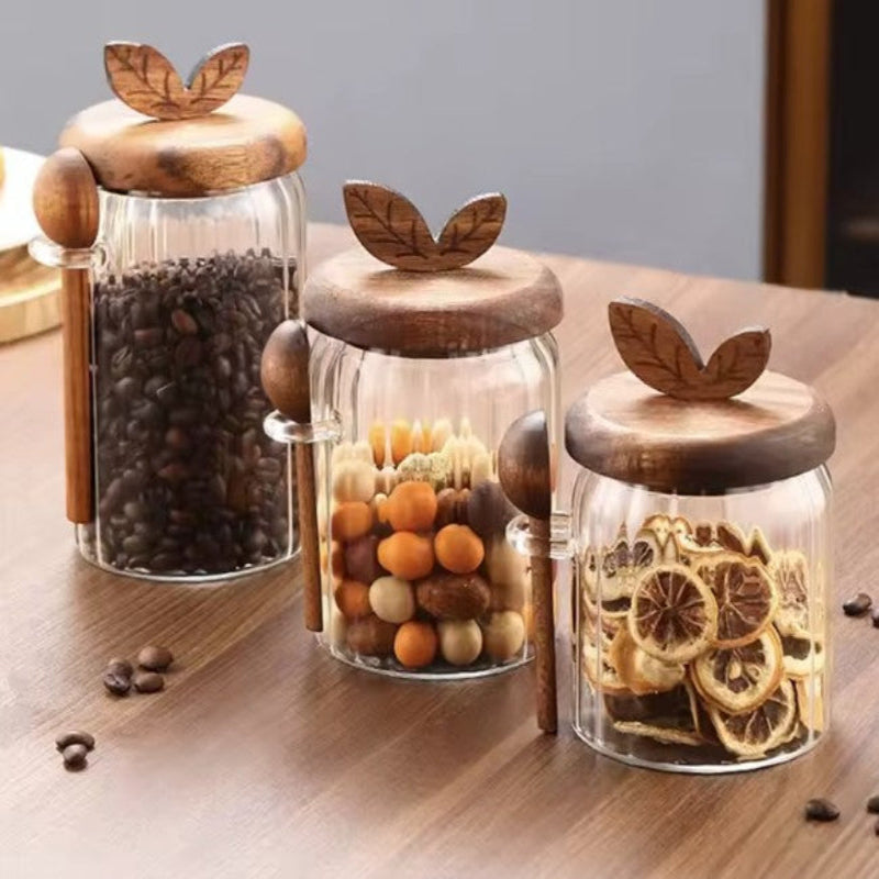 Three glass jars with wooden lids on a wooden surface, containing coffee beans, dried fruits, and citrus slices.