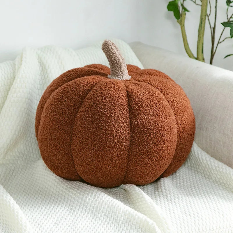 A soft, brown, pumpkin-shaped pillow with a textured stem rests on a white, woven blanket draped over a light-colored sofa.
