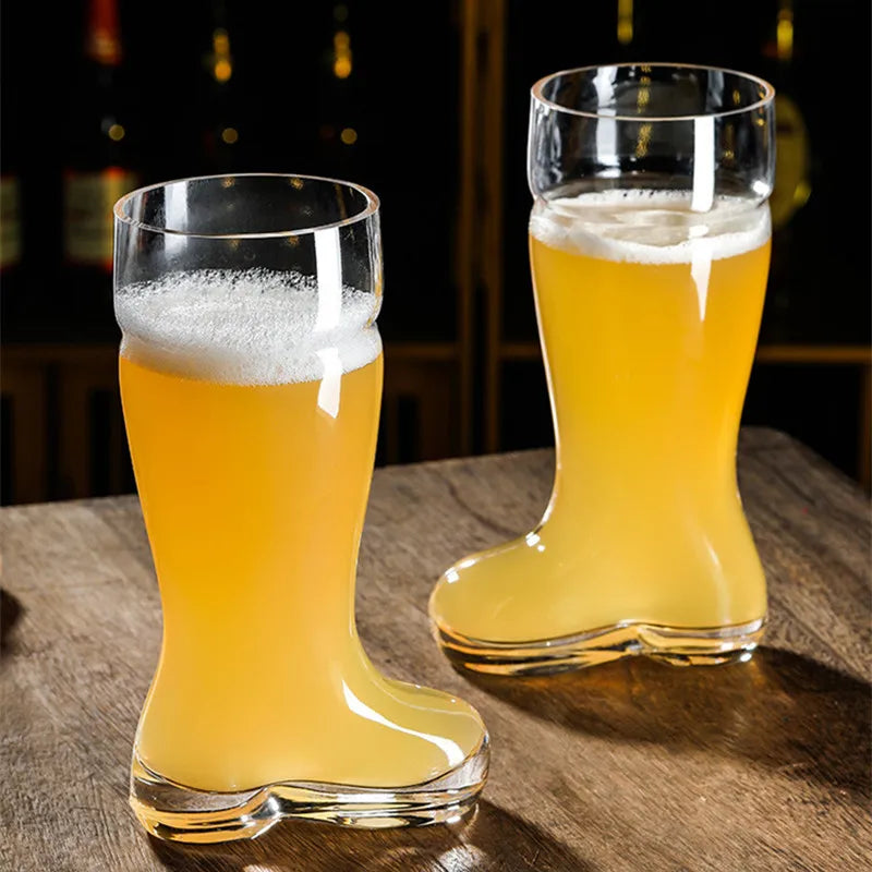 Two clear glass beer boot mugs, filled with light-colored beer and white foam, sitting on a wooden table in a dimly lit setting with blurred bottles in the background.