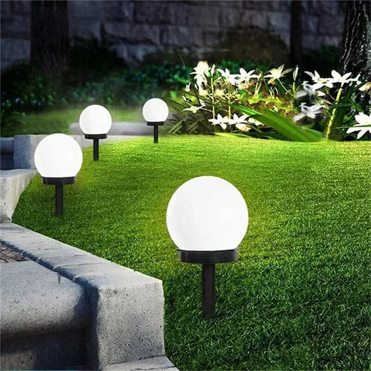 Four spherical white solar garden lights on black stakes illuminate a grassy area and a stone border at night. White flowers grow in the background.