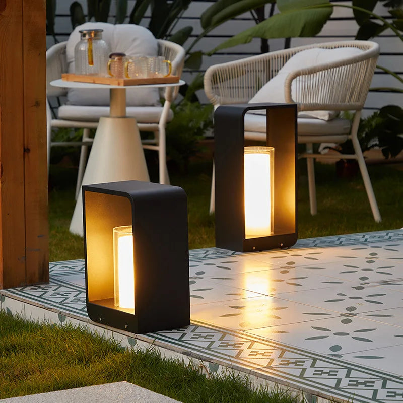 Two modern, dark rectangular outdoor pathway lights with glowing rectangular diffusers, illuminating a tiled patio and grassy area with outdoor furniture.