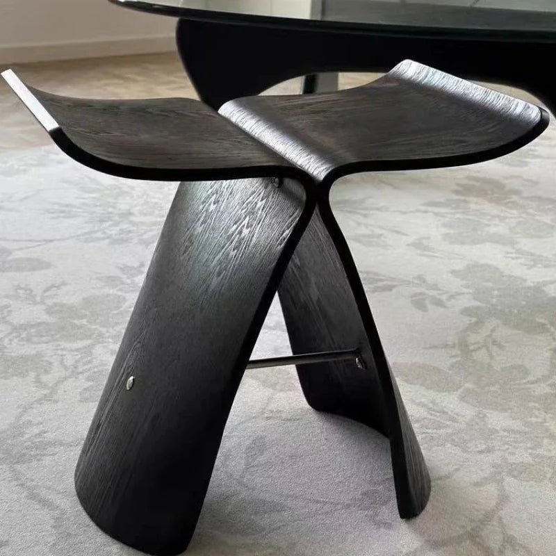 A black Hans Wegner Butterfly Stool, made of bent plywood, stands on a light gray patterned rug. Another piece of black furniture is partially visible in the blurred background.