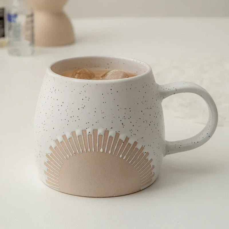 A ceramic mug with a speckled white glaze and an embossed sunburst design sits on a table, filled with an iced beverage. The curved handle and rustic aesthetic complement the minimalist background.