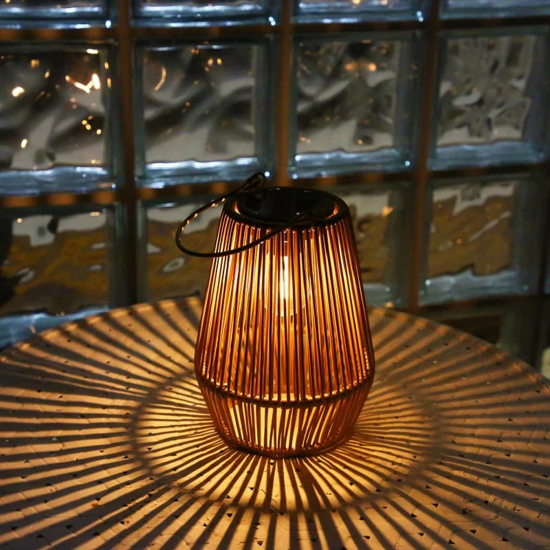 A warm-toned, cylindrical outdoor lantern made of vertical slats with a dark solar panel on top sits on a round, textured surface, casting radiating shadows. The lantern emits a soft glow and has a dark handle. A window with textured glass blocks is visible in the background.