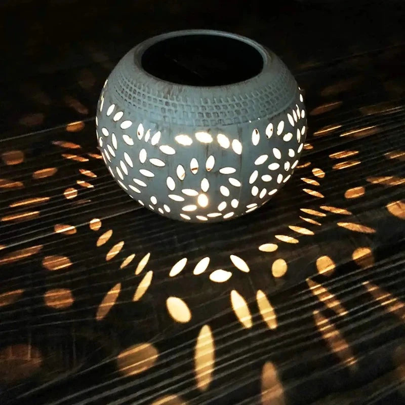 A round, off-white outdoor lantern with decorative oval cutouts emits a warm white glow, casting patterned shadows on a dark wooden surface at night. The lantern has a dark, flat top, likely a solar panel.