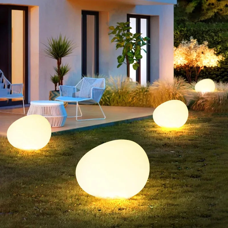A modern outdoor patio with white wicker furniture and round, glowing egg-shaped lights scattered on the green lawn at dusk.