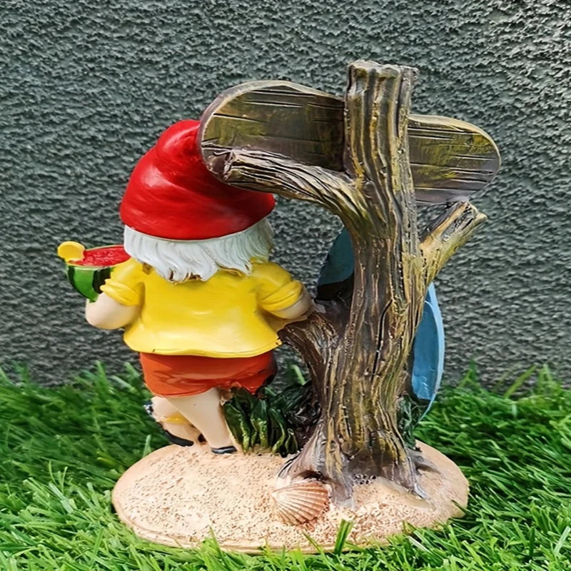 A garden gnome figurine is viewed from the back. It has a white beard and wears a red pointed hat, a yellow shirt, and red shorts with black shoes. The gnome holds a slice of watermelon. Behind the gnome is a rustic wooden sign attached to a tree-like structure, leaning against a blue surfboard. The figurine stands on a sandy base with seashells, placed on artificial green grass. The background is a textured grey wall.