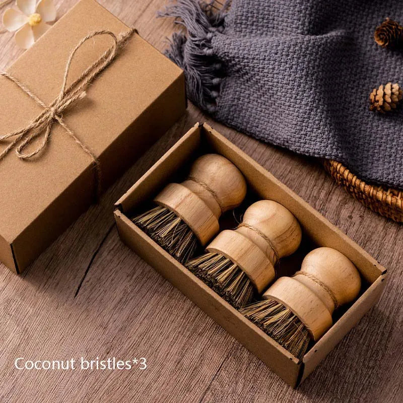 Set of three wooden scrub brushes with coconut fiber bristles in a kraft paper gift box tied with twine. Placed on a rustic wooden surface with a textured fabric, pinecones, and decorative elements in the background.