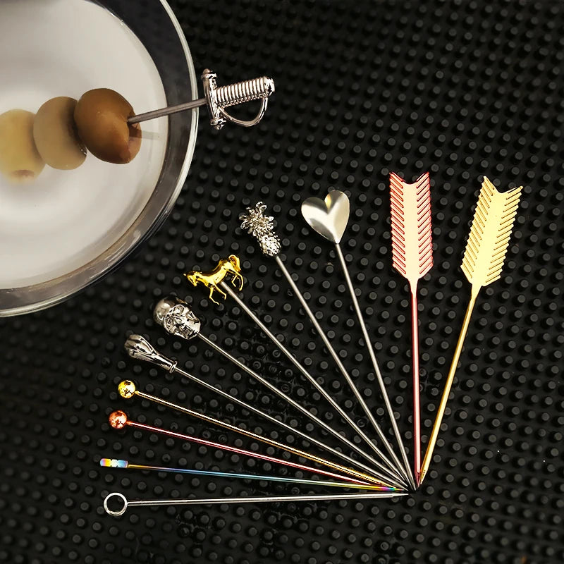 A variety of decorative stainless steel cocktail picks arranged in a fan shape on a black textured surface, with a plate of olives in the upper left corner.