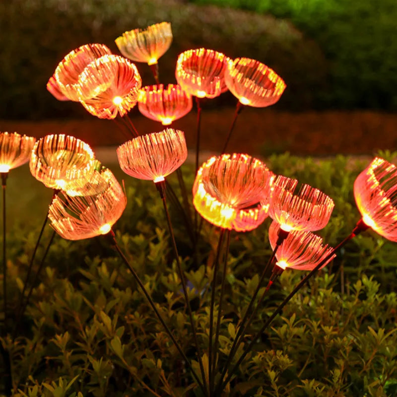 Numerous solar-powered garden lights with a jellyfish-like appearance emit a warm orange glow in a garden at night, standing above dark green foliage.