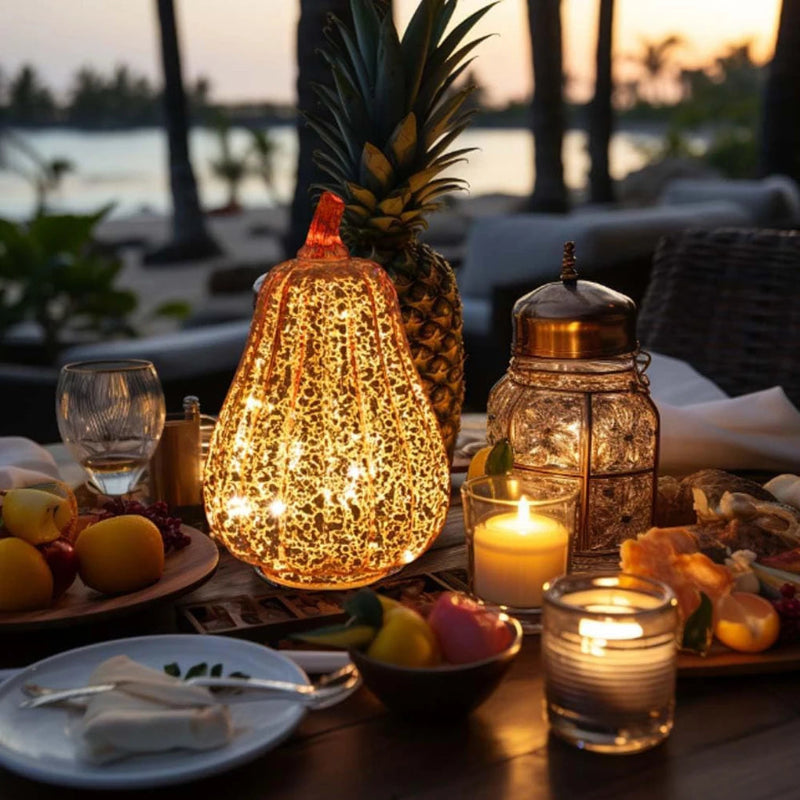 A pear-shaped, glowing, gold pumpkin decoration on a dining table with a pineapple, candles, and food.