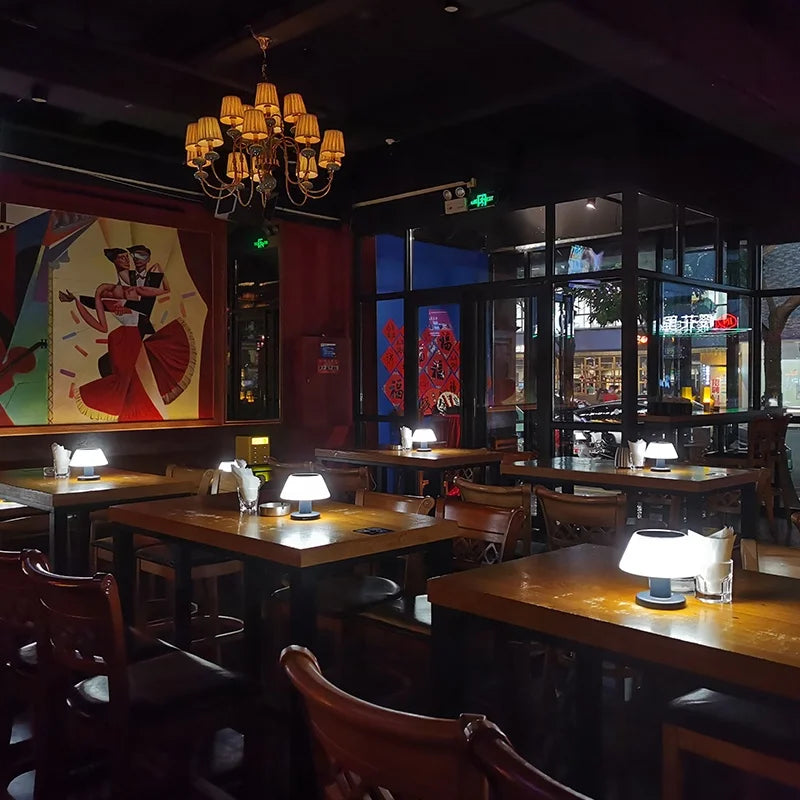 Several small, modern table lamps with white shades illuminate wooden tables in a dimly lit restaurant or bar. Each table is set with napkins and some have glasses. A large, colorful abstract painting hangs on a red wall in the background, and a multi-tiered chandelier hangs from the dark ceiling. Large windows show the outside night scene. Wooden chairs are placed around the tables.