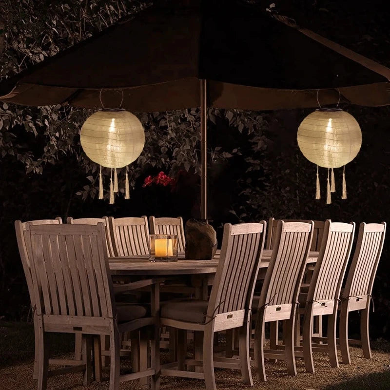 Two round, white solar lanterns with tassels hang beneath a dark patio umbrella, illuminating a wooden dining table set with lit candles.