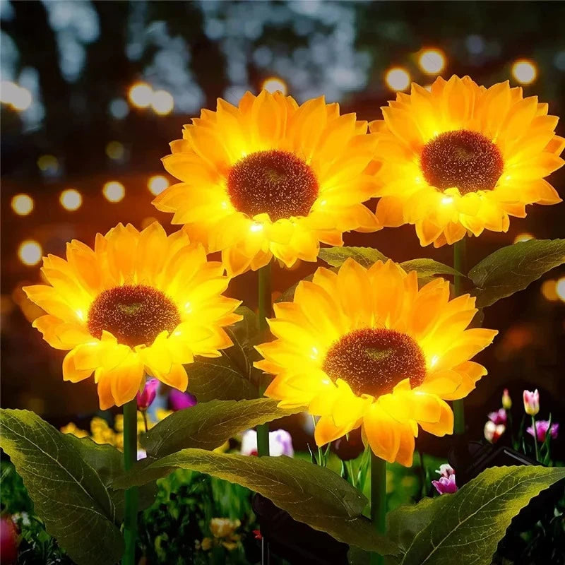 Four artificial sunflowers with brown centers and bright yellow glowing petals are illuminated in a garden at night. The flowers stand on green stems with leaves, and warm white bokeh lights are visible in the blurred background, suggesting a festive or magical atmosphere.