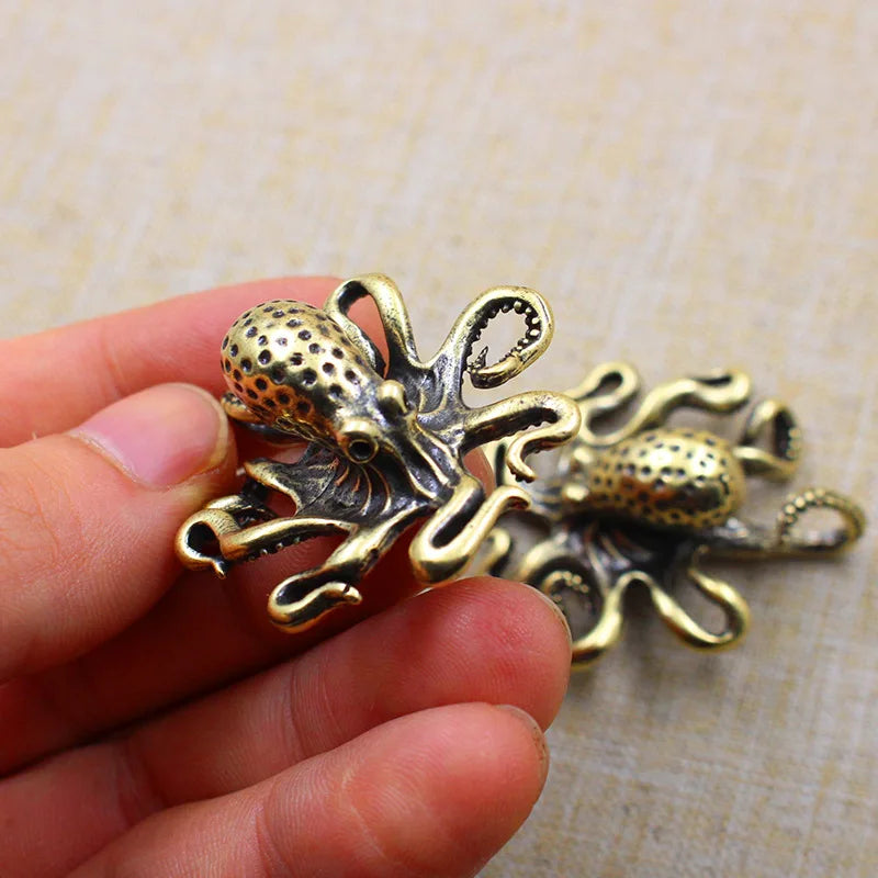 A close-up image of a small metallic octopus-shaped object with an antique brass or bronze finish, held between a person's fingers. The octopus has detailed tentacles and a textured head. Another identical octopus figurine is resting on a neutral fabric surface in the background. The objects appear to be decorative, possibly used as charms, figurines, or accessories.