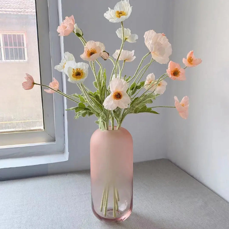 A frosted pink vase holds a bouquet of white and pink poppies by a window.