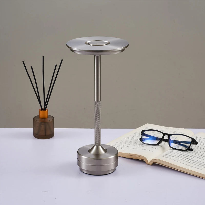 Modern silver table lamp with a sleek, disc-shaped LED light source and a slim, metallic stem with a textured grip. Placed on a white desk alongside an open book, reading glasses, and a reed diffuser in a minimalist setting.