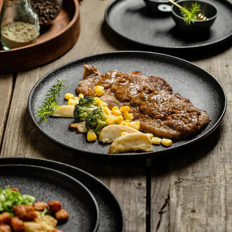 A round, matte black plate with a steak and vegetables on it, placed on a rustic wooden table.