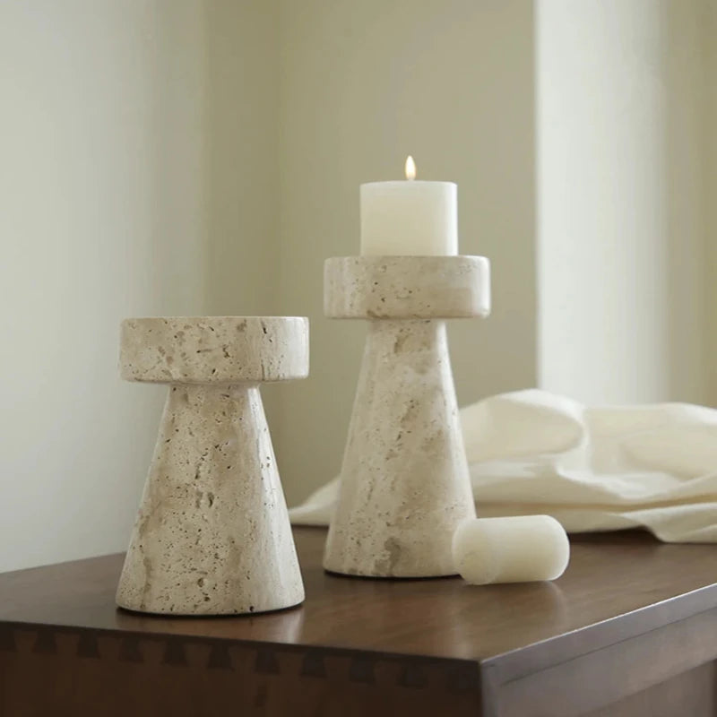 Two textured, light-colored faux travertine stone candle holders of different heights, one holding a lit white pillar candle, arranged on a dark wooden surface with an unlit white pillar candle and a soft white cloth.