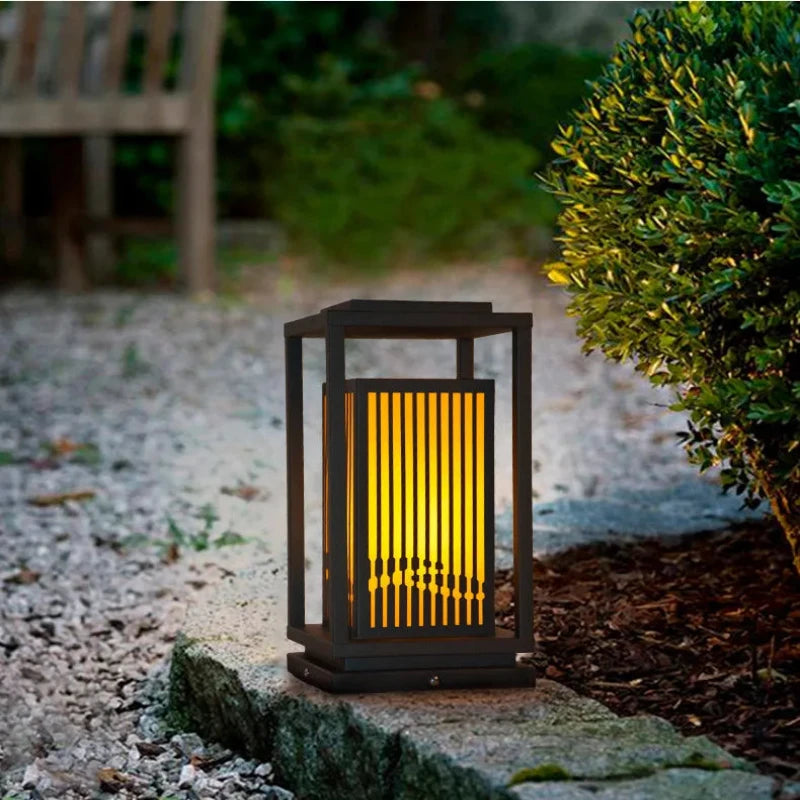 A modern, dark-framed outdoor lantern with vertical slats emits a warm yellow glow as it stands on a weathered stone in a garden setting with small pebbles and greenery.