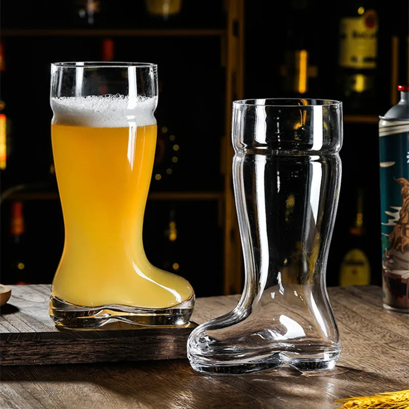 Two clear glass beer boot mugs, one filled with a light-colored beer and white foam, sitting on a dark wooden surface against a blurred background of a bar shelf with various bottles.