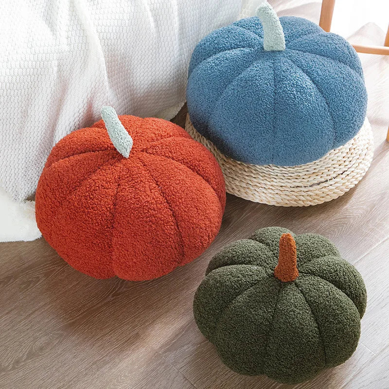 Three textured pumpkin-shaped pillows in blue, orange, and green are arranged on a light-colored floor, with the blue one resting on a woven mat.
