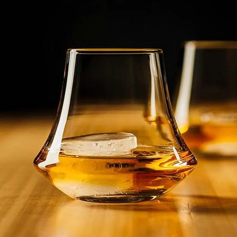 A modern whiskey glass with a unique curved design, filled with golden whiskey and a single ice cube, set on a warm wooden surface with another glass slightly blurred in the background.