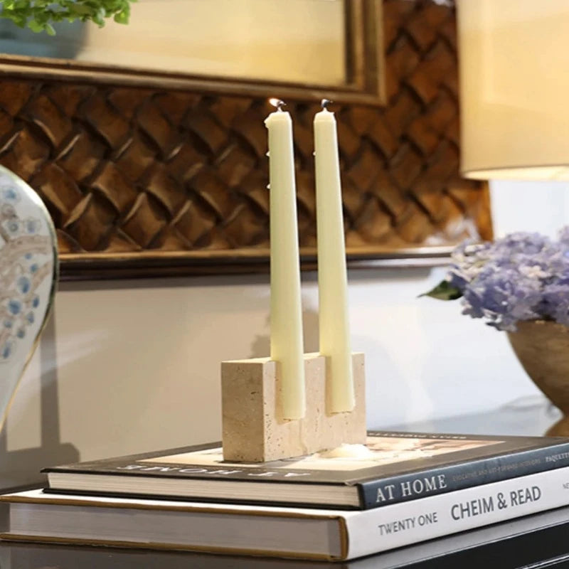 Two lit, pale yellow taper candles in a textured, light-colored travertine stone block holder, resting on a stack of books on a dark surface, with blurred decorative objects in the background.