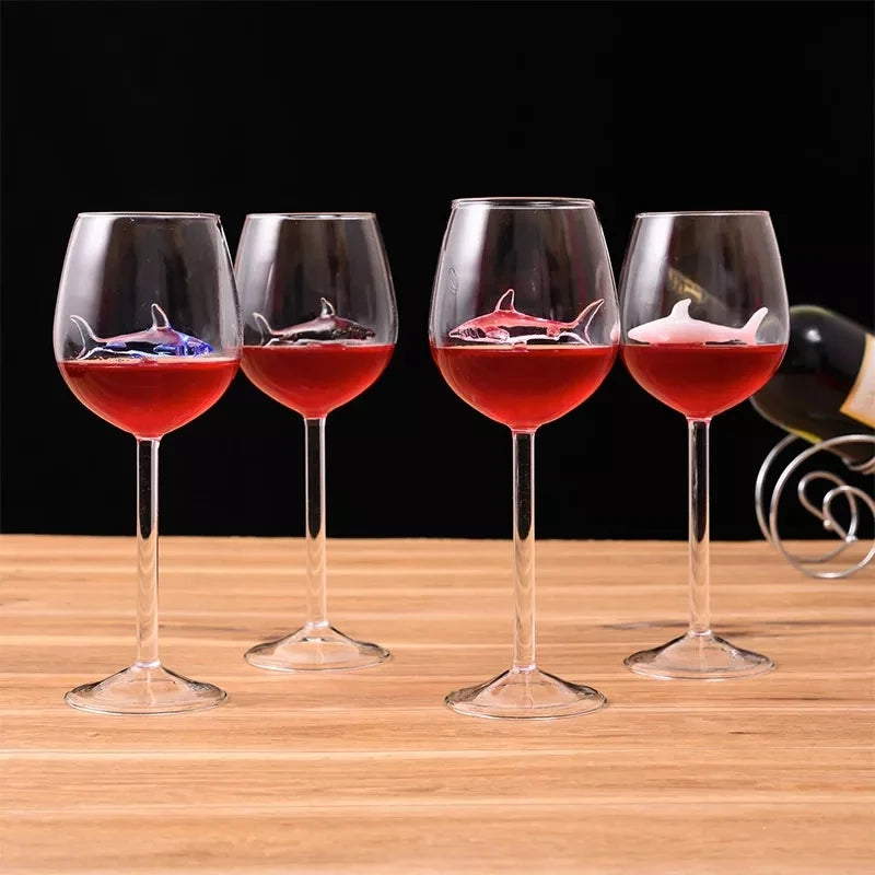 A set of four clear wine glasses, each containing a small, colored glass shark figurine suspended inside, filled with a red liquid. A bottle of wine is visible in the background.