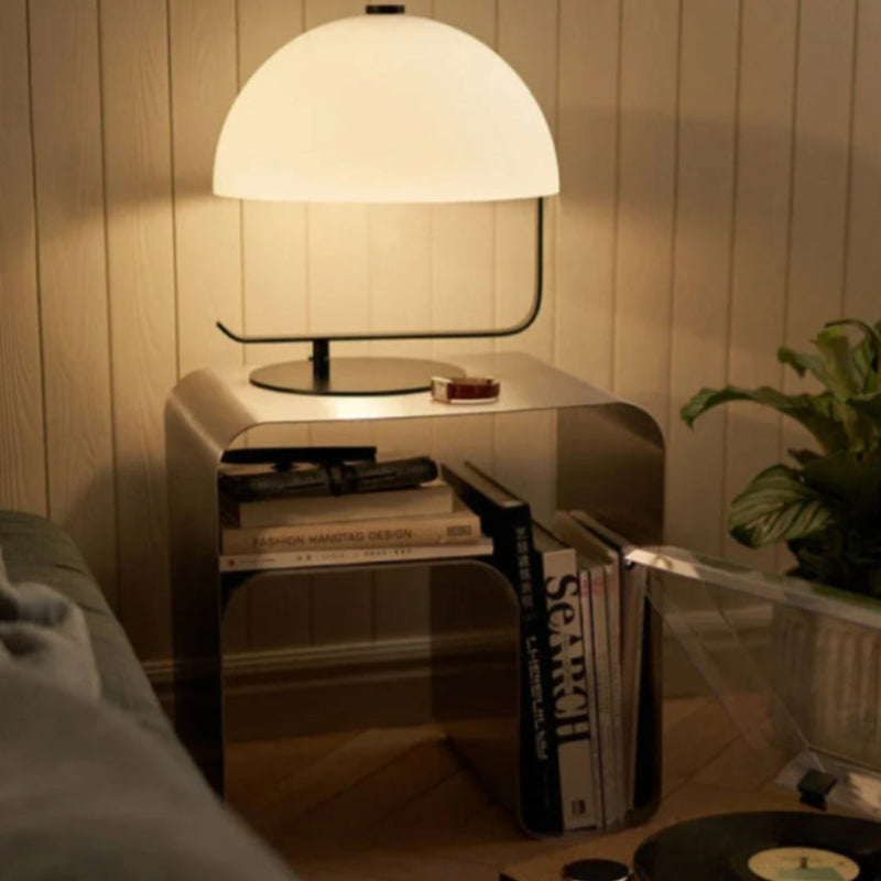 A white, half-dome shaped lamp illuminates a silver metal nightstand with books and a plant.
