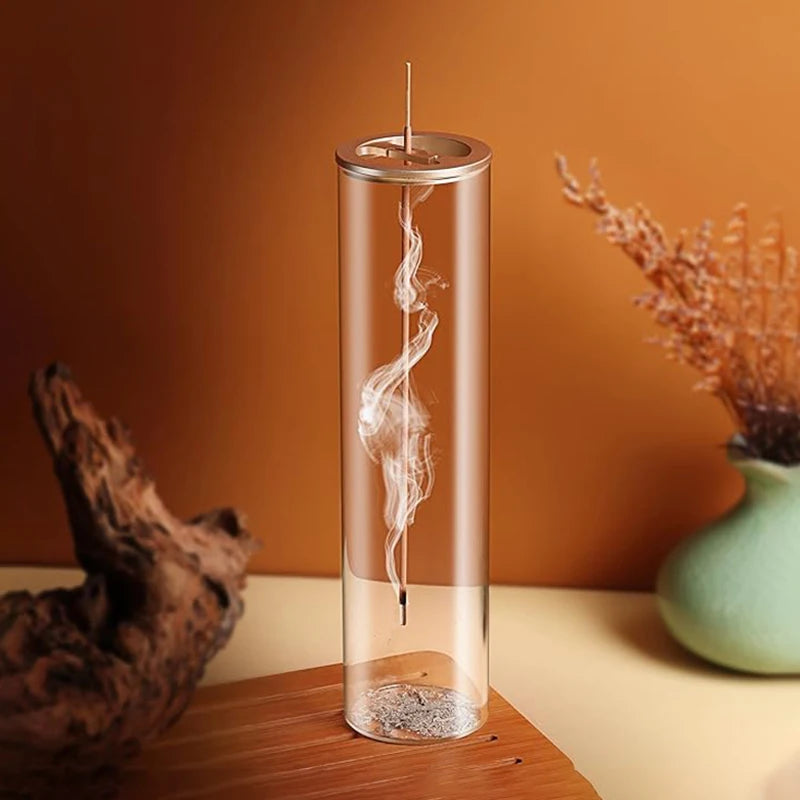 This image features a modern and elegant incense burner. The incense stick is suspended within a tall, transparent glass cylinder, allowing the smoke to swirl beautifully as it rises. The base collects the ash, keeping the setup clean and stylish. The background has warm earthy tones with a wooden surface, a decorative vase with dried plants, and a natural wooden accent, adding to the aesthetic and tranquil ambiance.