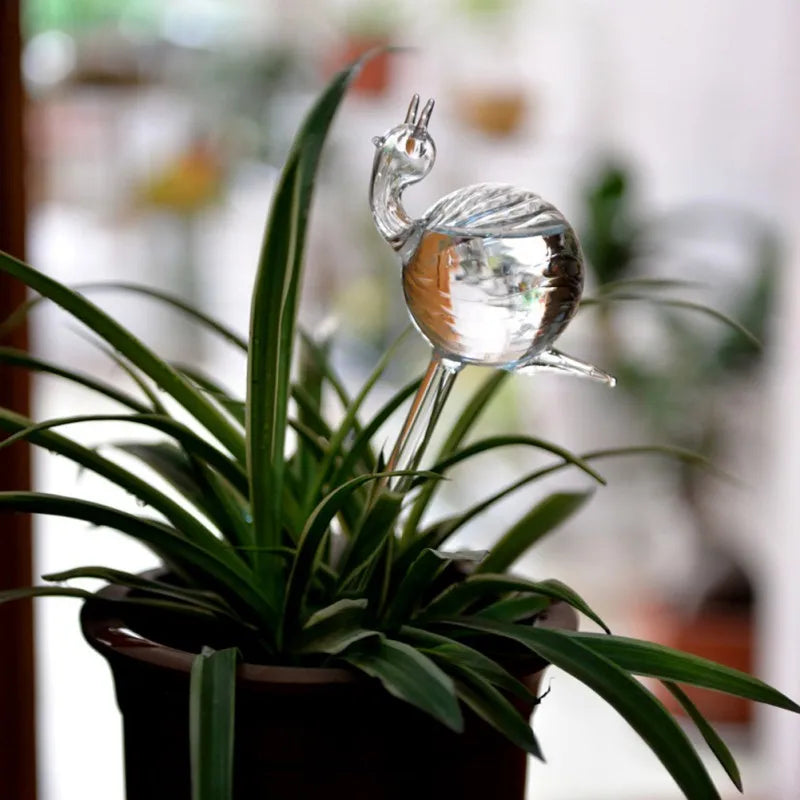 A plant in a pot has a clear, hand-blown glass snail-shaped self-watering device inserted into the soil.