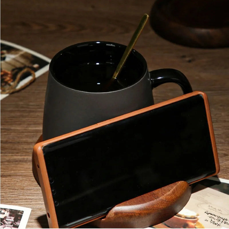 A black mug with a gold spoon, a brown smartphone in a wooden stand, and old photographs on a wooden table.