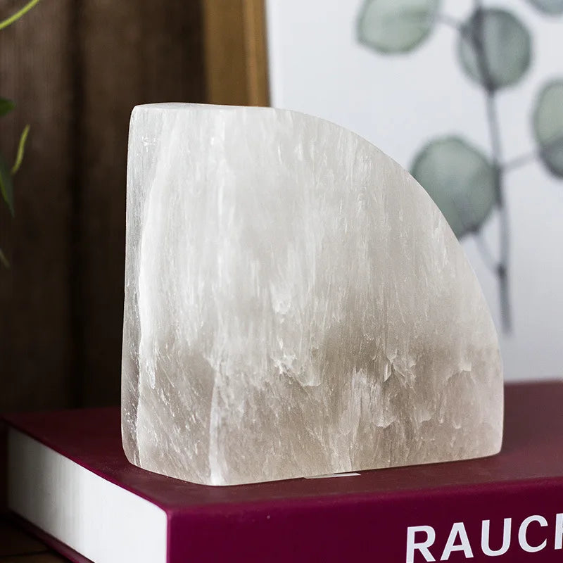 A close-up of a white crystal bookend with a smooth, semi-circular design, placed on a red hardcover book, with a blurred botanical print in the background.