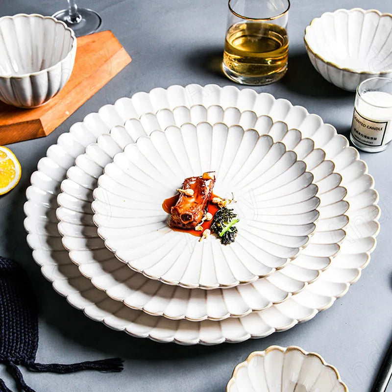 A place setting of white ceramic scalloped-edge plates with raised ridges, with a main course plate on top, next to a bowl, a glass, and a candle.