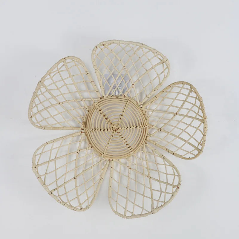 A decorative woven rattan flower with an openwork lattice petal design. The handcrafted wall decor piece features a natural beige color, a circular woven center, and five delicate petals made of intersecting rattan strands. Perfect for bohemian or rustic home decor.
