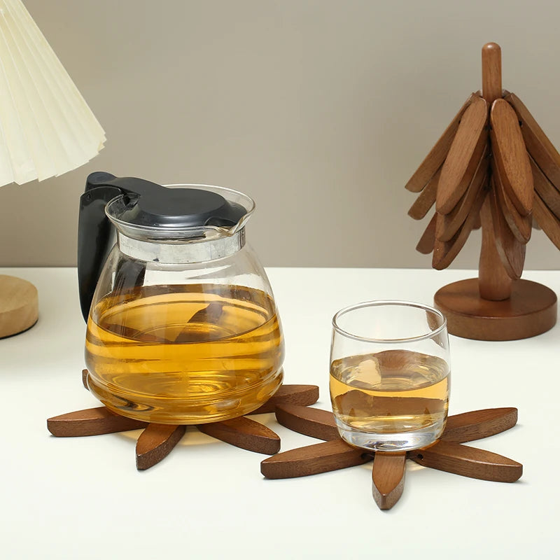 The image shows three wooden coasters: two tree-shaped and one starburst-shaped. A glass and a teapot are on the starburst coaster, and a tree-shaped coaster stands upright.
