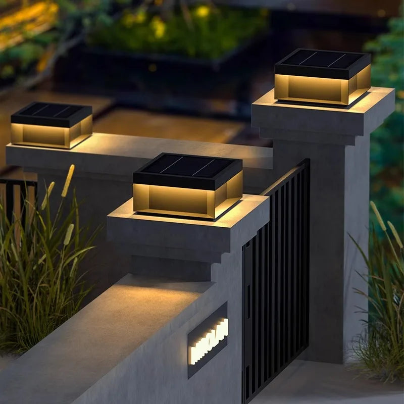 A square, modern, waterproof, solar-powered outdoor wall lamp with a dark top and glowing warm-toned panels, mounted on a gray concrete pillar.