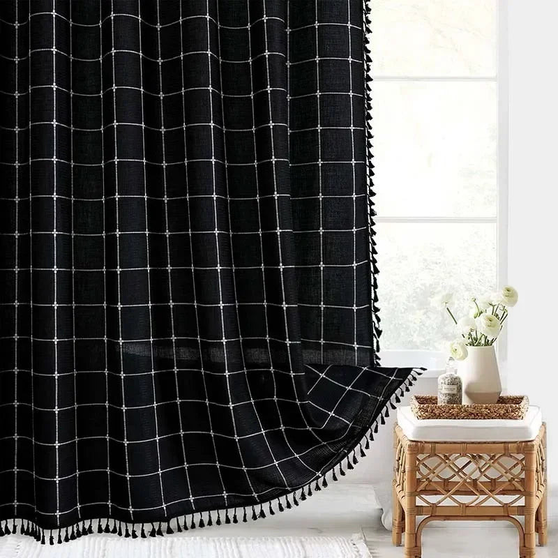 Black grid curtain with tassels in a room with a wooden side table and white flowers.