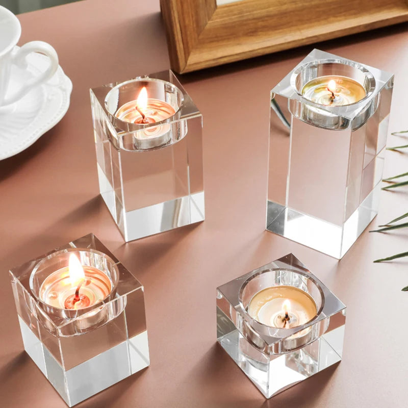 Four clear, square crystal candle holders, each containing a lit tea light candle, are arranged on a light brown surface. Two are shorter and two are taller. A white teacup and saucer are partially visible in the upper left corner, and a wooden frame and some green foliage are in the blurred background. The candles cast a warm glow.