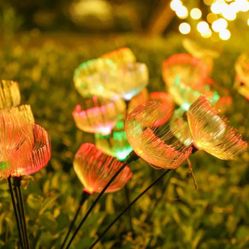 Several solar-powered garden lights with a jellyfish-like shape emit a blend of orange, yellow, and green glows in a garden at night, positioned above dark foliage. Blurred string lights are visible in the upper background.