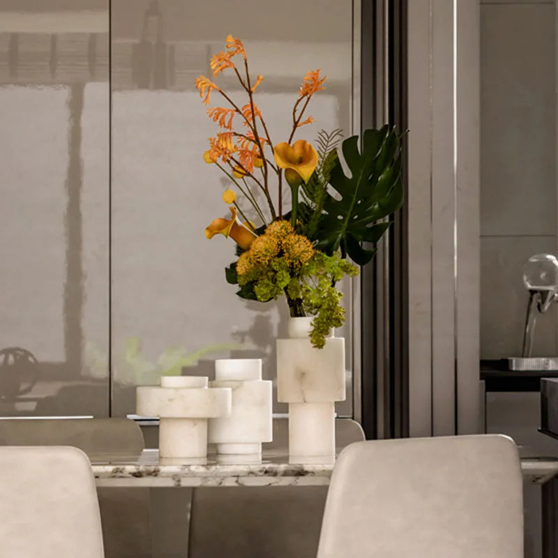 A dining table adorned with Scandinavian White Onyx vases, featuring a modern geometric design. One vase holds a vibrant floral arrangement with orange and green foliage, adding a touch of nature to the elegant setting. The background showcases a contemporary interior with neutral tones and reflective surfaces.