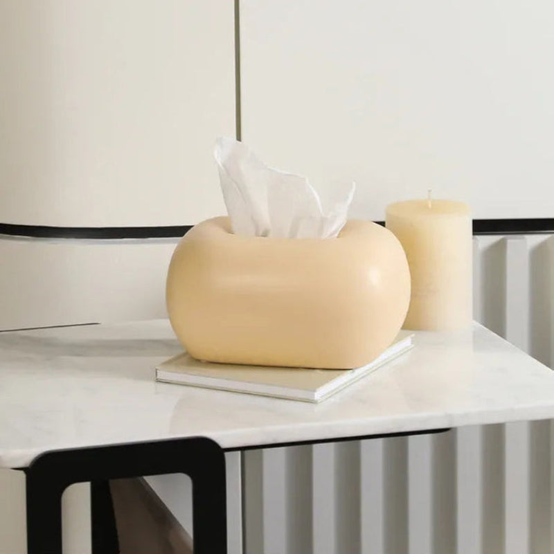 A light yellow, donut-shaped tissue box holder sits on a white marble surface next to a cream-colored candle.