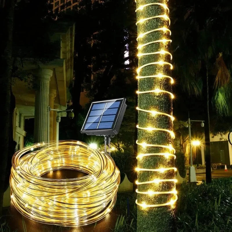 A long strand of warm white LED fairy lights is tightly coiled on the ground next to a small solar panel on a stake. The same string of lights is wrapped around the trunk of a nearby tree, illuminating it in a spiral pattern at night. A building and other outdoor lights are visible in the blurred background.